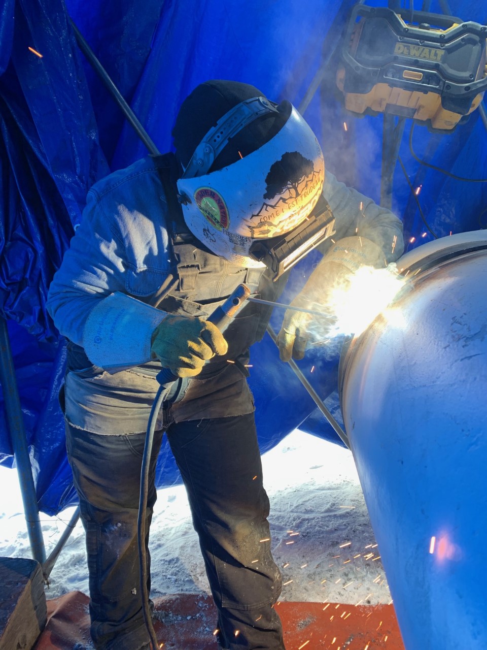 Joelle Milne welding a pipe. She wears full PPE including hood. Blue tent is in the background.